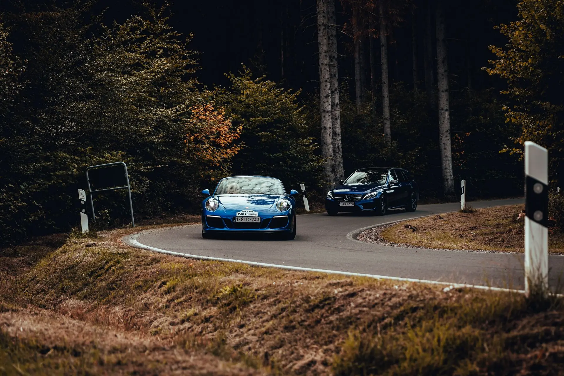 Blue Porsche 911 vs Mercedes-Benz C63 AMG driving a windy road in the woods